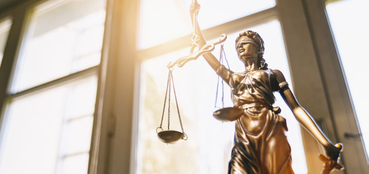 Bronze statue of Lady Justice holding a balance scale and sword stands in front of a large sunlit window, symbolizing fairness and law. The figure is blindfolded, representing impartiality.