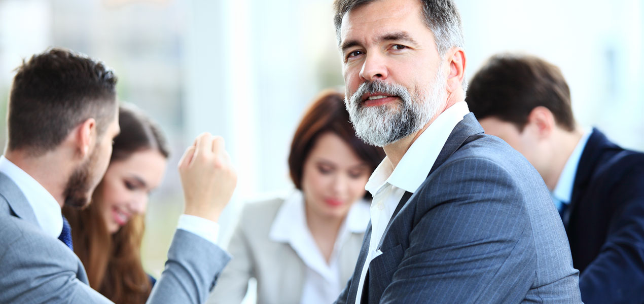 A group of business people sitting around a table chatting, with one man turned around smiling into the camera.